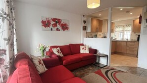 Een woonkamer met een rode bank, een kleine tafel en een bloemenvaas. Achtergrond toont een keuken.