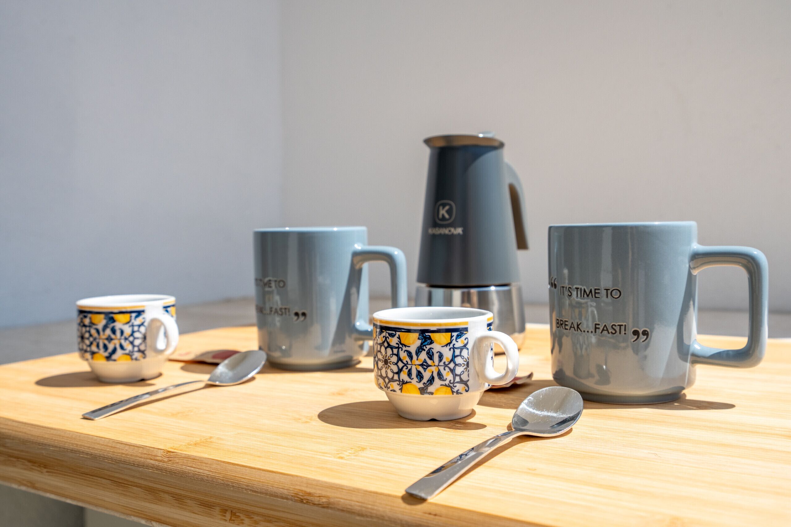 A wooden table with four ceramic mugs, a spoon, and a coffee maker.