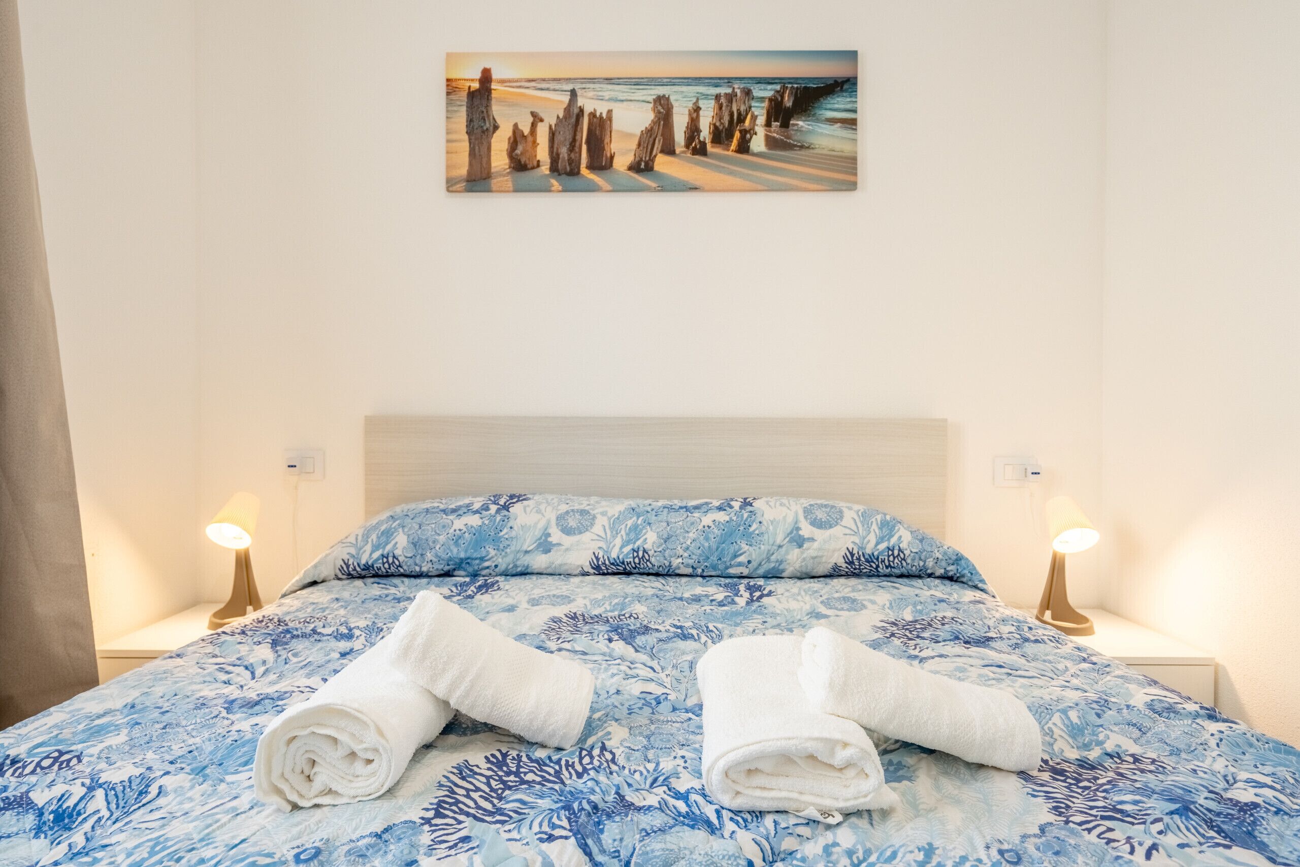 A bed with a floral bedspread, two rolled towels, bedside lamps, and a painting of a beach scene.