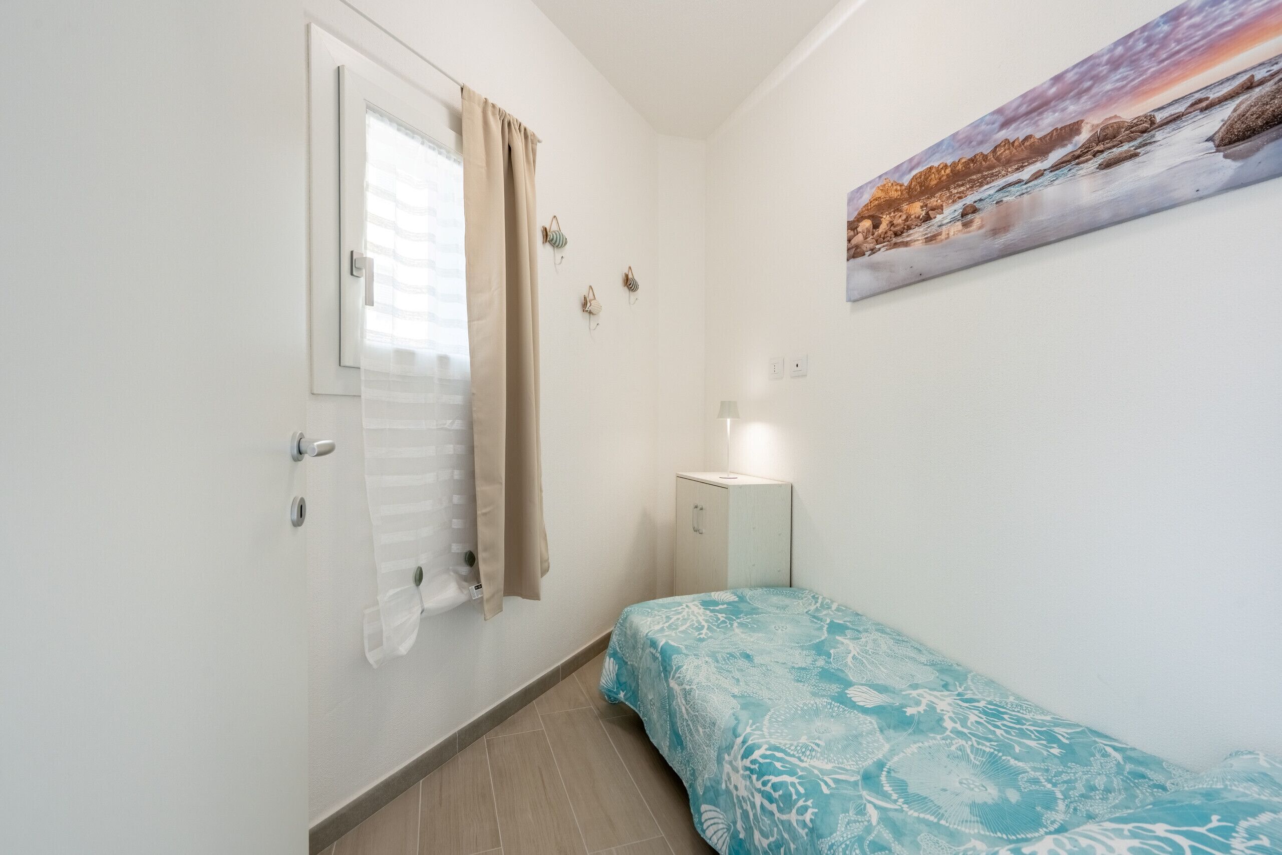 Ein kleines Schlafzimmer mit Bett, Tür, Fenster mit Vorhängen, Nachttisch mit Lampe und Wandbild.