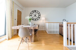 A dining area with a wooden table, chairs, a large wall clock, a vase with flowers, and a lamp. - 3 Bed in Bideford (oc-52079) (Westward Ho!)