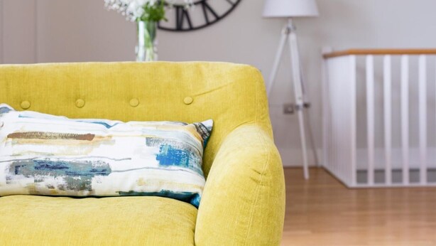 Ein gelbes Sofa mit farbigem Kissen, eine Wanduhr, eine Lampe und ein Kinderbett in einem Raum mit Holzboden.