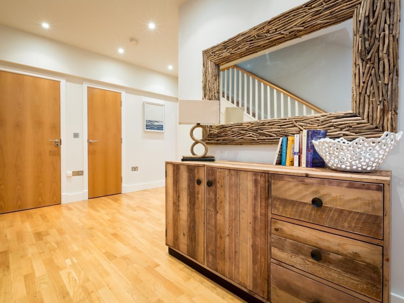 Una habitación con piso de madera, un mueble de madera con cajones, un espejo con marco tejido, una escalera con barandales blancos y un cuadro enmarcado en la pared.