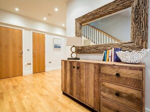 A room with wooden flooring, a wooden cabinet with drawers, a mirror with a woven frame, a staircase with white railings, and a framed picture on the wall. - 3 Bed in Bideford (oc-52079) (Westward Ho!)