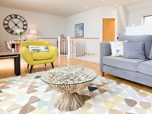 A living room with a yellow armchair, a grey sofa, a glass coffee table with a woven base, a large wall clock, and a painting. - 3 Bed in Bideford (oc-52079) (Westward Ho!)