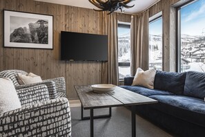 Ein modernes Wohnzimmer mit einer Couch, einem gemusterten Sessel, einem Holztisch, einem Flachbildfernseher und einem großen Fenster mit Bergblick.