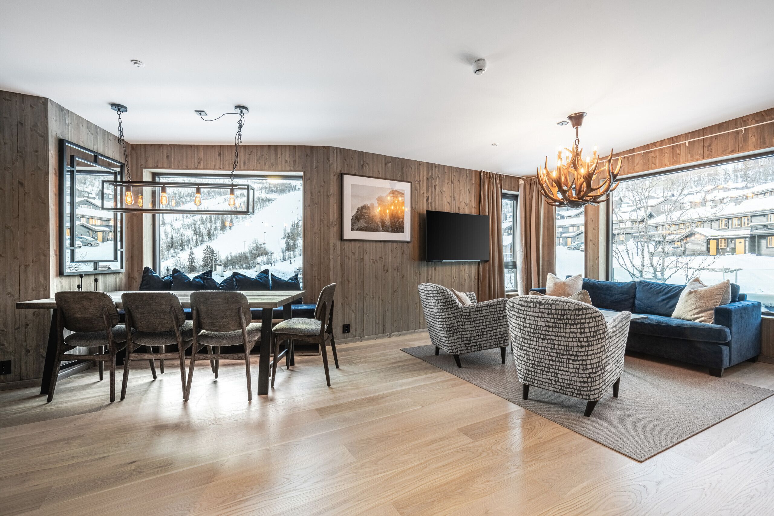 Un salon moderne comprenant un coin repas, des murs en bois, de grandes fenêtres, un lustre et un canapé bleu.