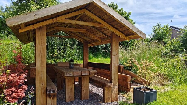 Ein hölzernes Pavillon mit einer Bank und einem Tisch, umgeben von Kies und Pflanzen.