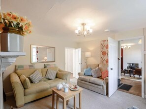 Ein Wohnzimmer mit zwei Sofas, einem Couchtisch, einer Blumenvase, einer Chandelierleuchtre und einem Kamin im Hintergrund.