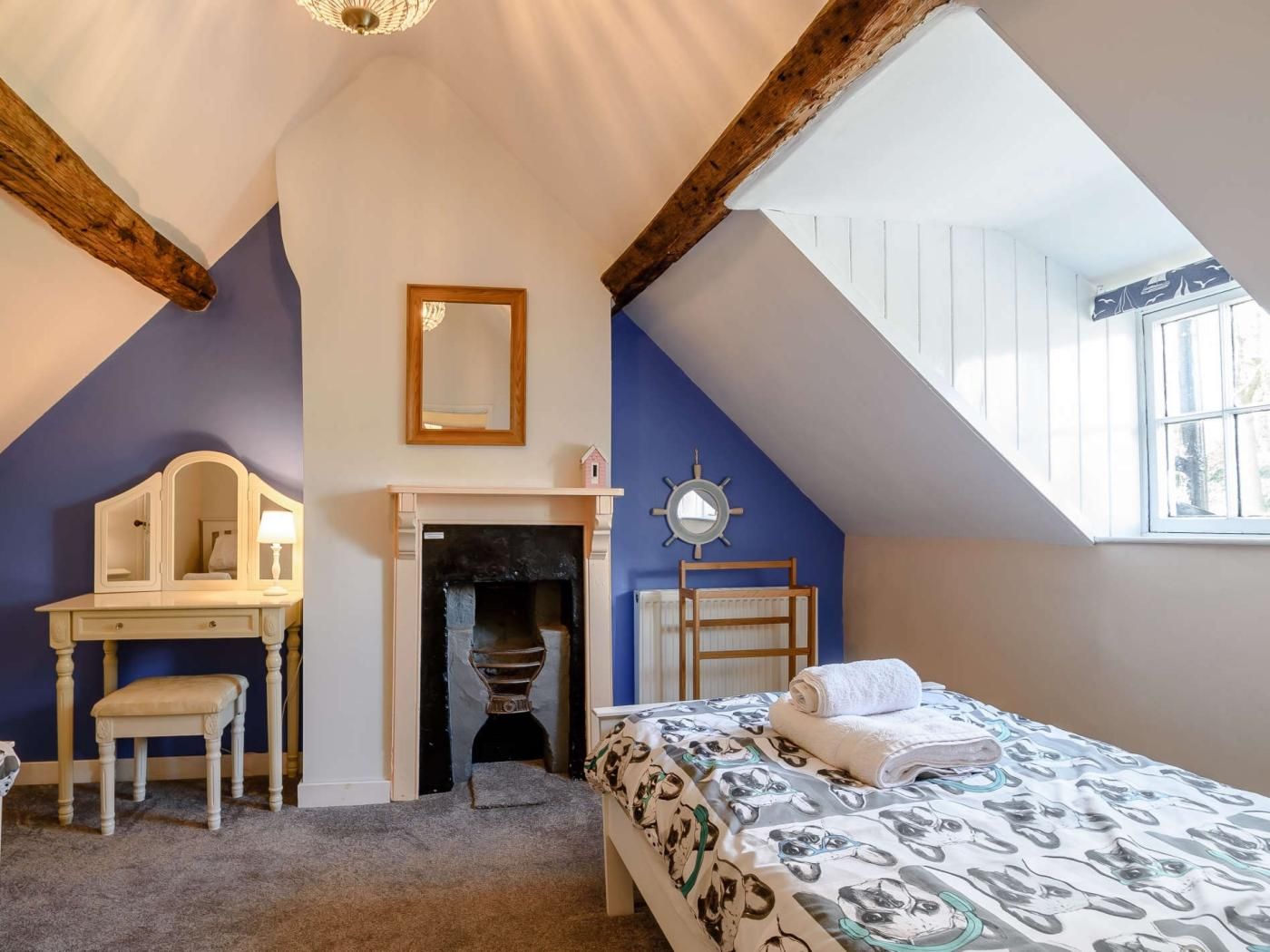 Une chambre avec un plafond en pente, une cheminée, un lit recouvert d’une couette à motifs, un bureau avec une chaise et une fenêtre donnant sur un beau paysage.