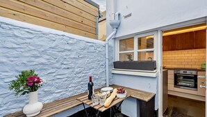 Un pequeño comedor al aire libre con un banco de madera, una mesa con comida y bebidas, una botella de vino y vistas a una cocina con pared de ladrillo y horno.