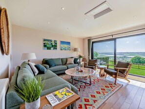 A modern living room with a sofa, chairs, a coffee table, and a large window offering a view of the countryside. - 3 Bed in Barnstaple (oc-53109) (Braunton)