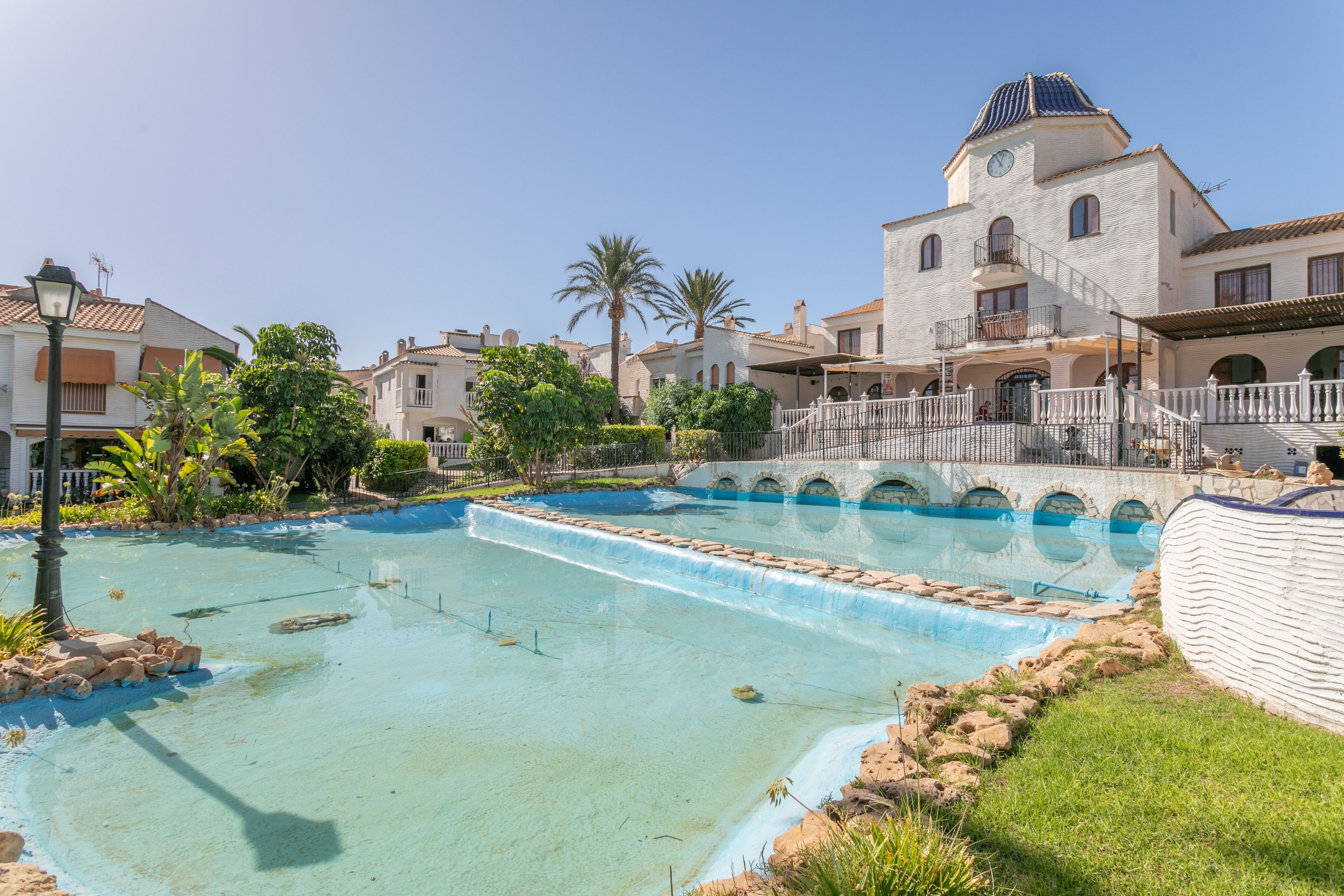 A swimming pool with a fountain, surrounded by buildings and palm trees.
