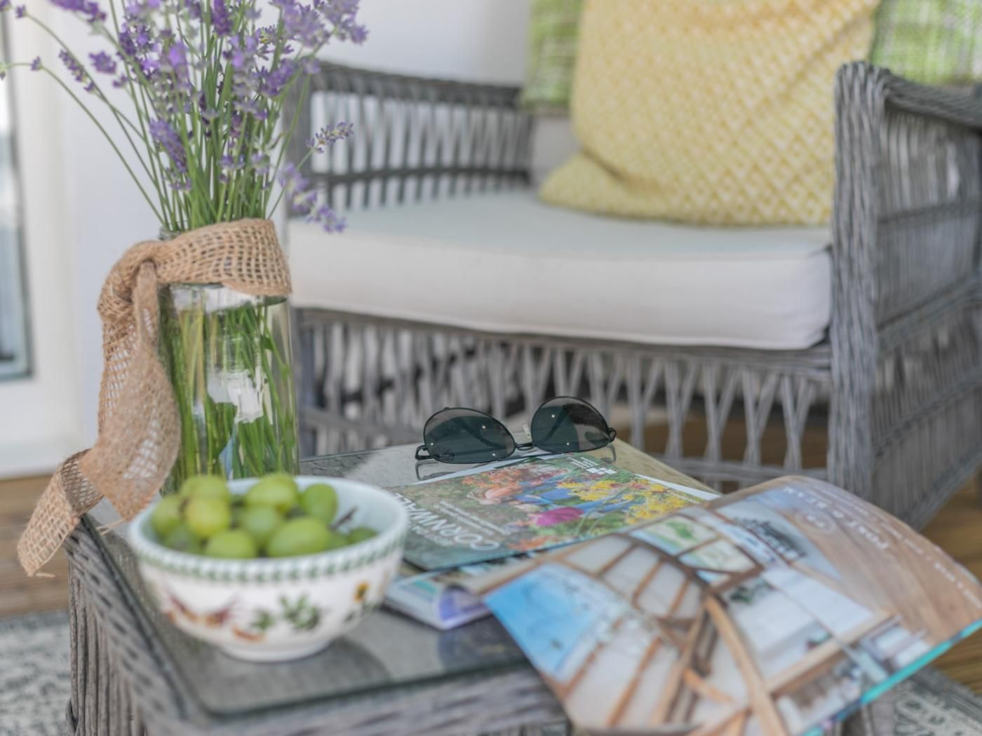 A wicker chair with a cushion, a glass vase with purple flowers, a bowl of green apples, a pair of sunglasses, and a magazine on a glass table.