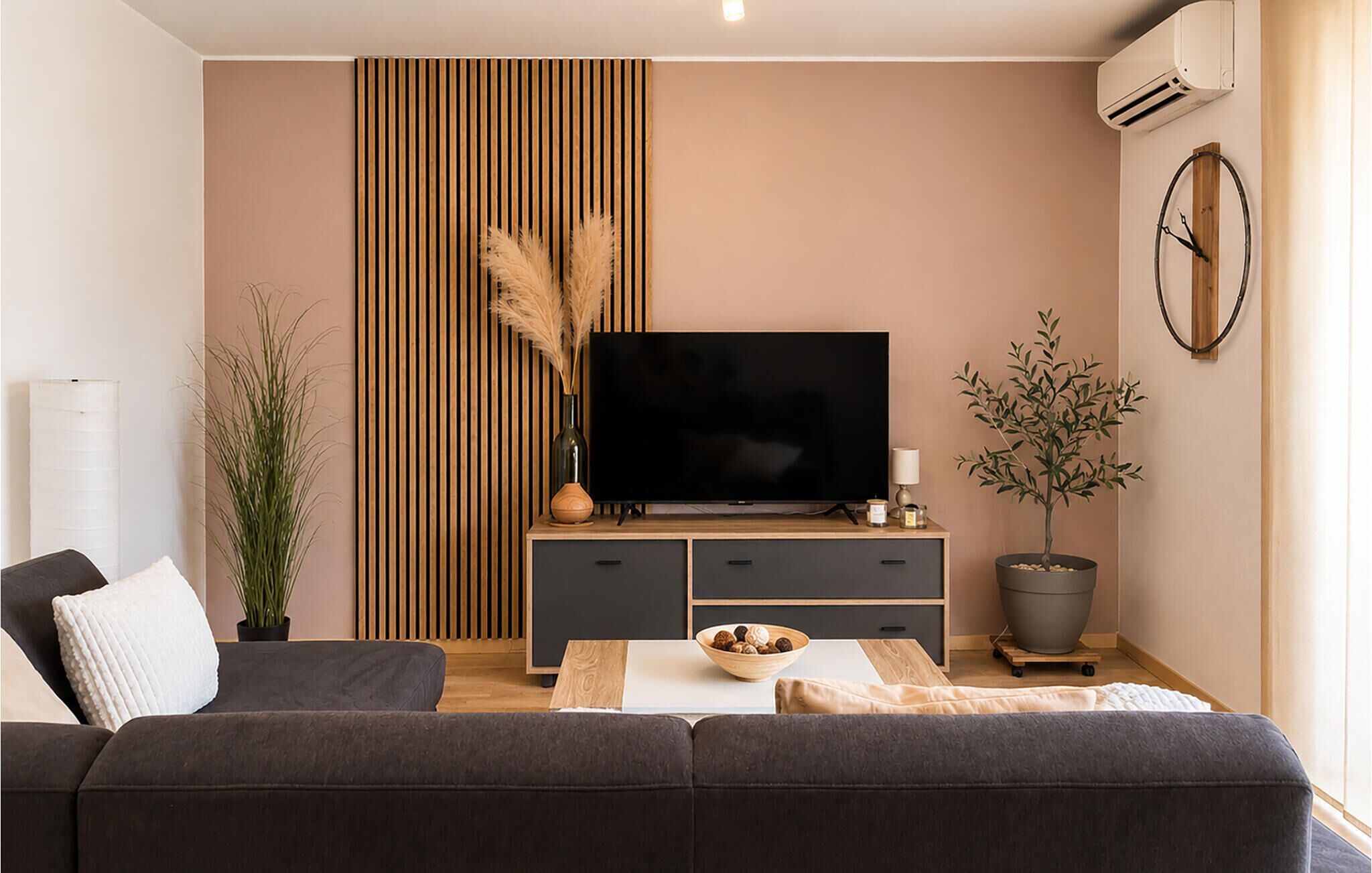 A modern living room with a sofa, a TV on a wooden stand, a potted plant, and a wall-mounted air conditioner.