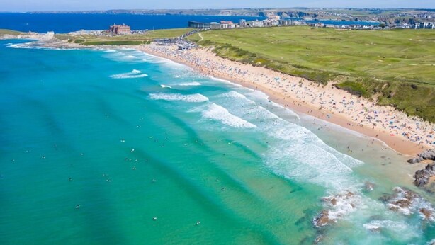 An aerial view of a coastal beach with clear waters, a sandy shore, and a distant building complex. - 3 Bed in Newquay (oc-53688) (Newquay)