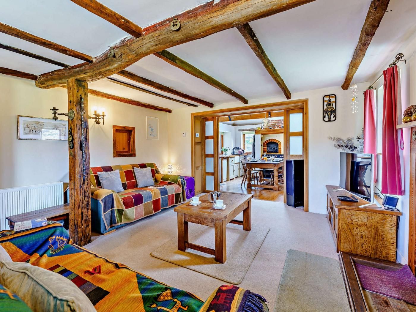 A cozy living room with exposed wooden beams, a colorful sofa, a wooden coffee table, and a kitchen area visible through an open doorway.