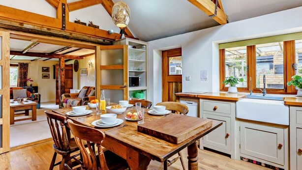 Une cuisine rustique avec des poutres en bois, une table à manger dressée pour le petit-déjeuner et une vue sur l’extérieur par une fenêtre.