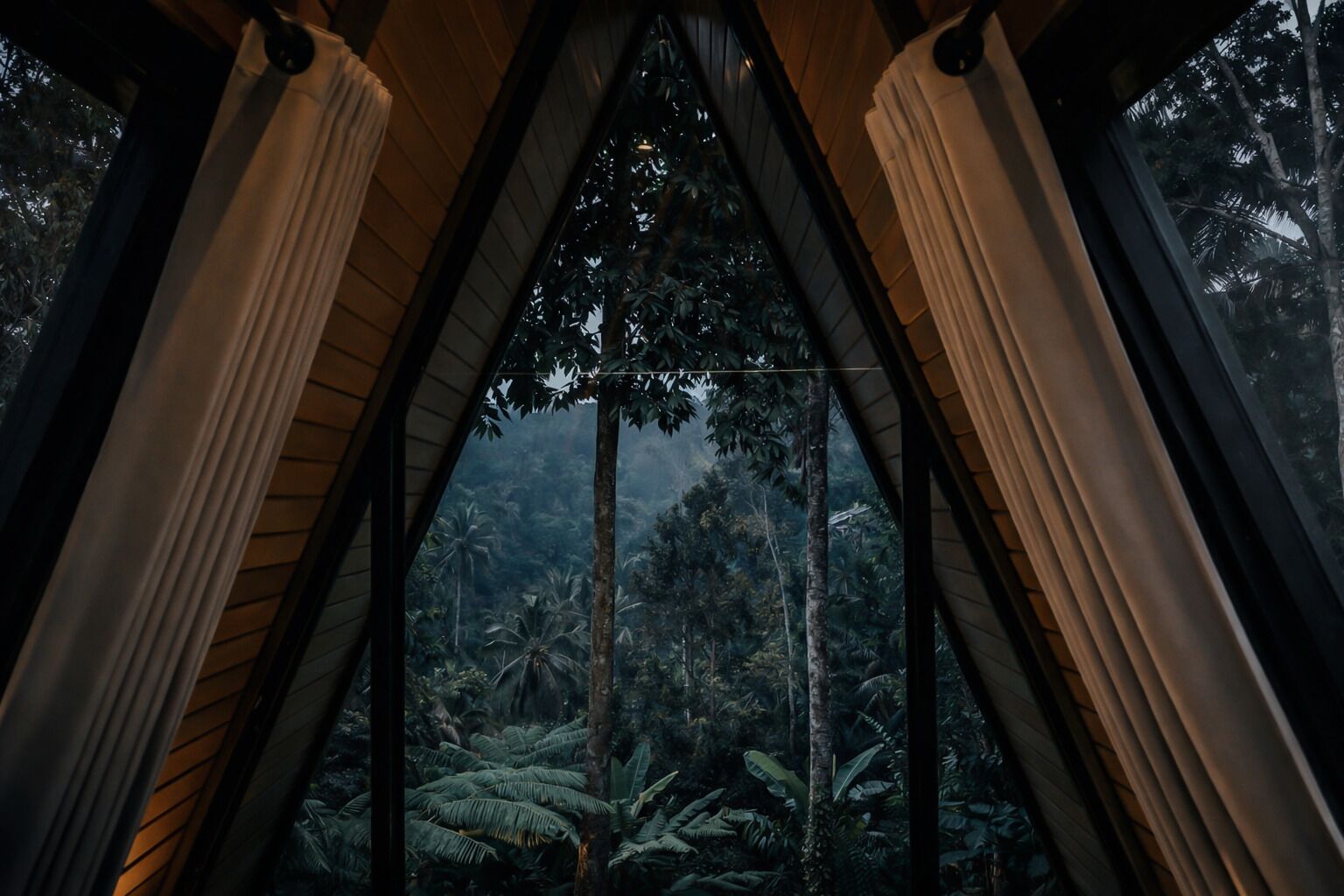 Una habitación con ventanal triangular, cortinas y vistas a densa vegetación tropical.