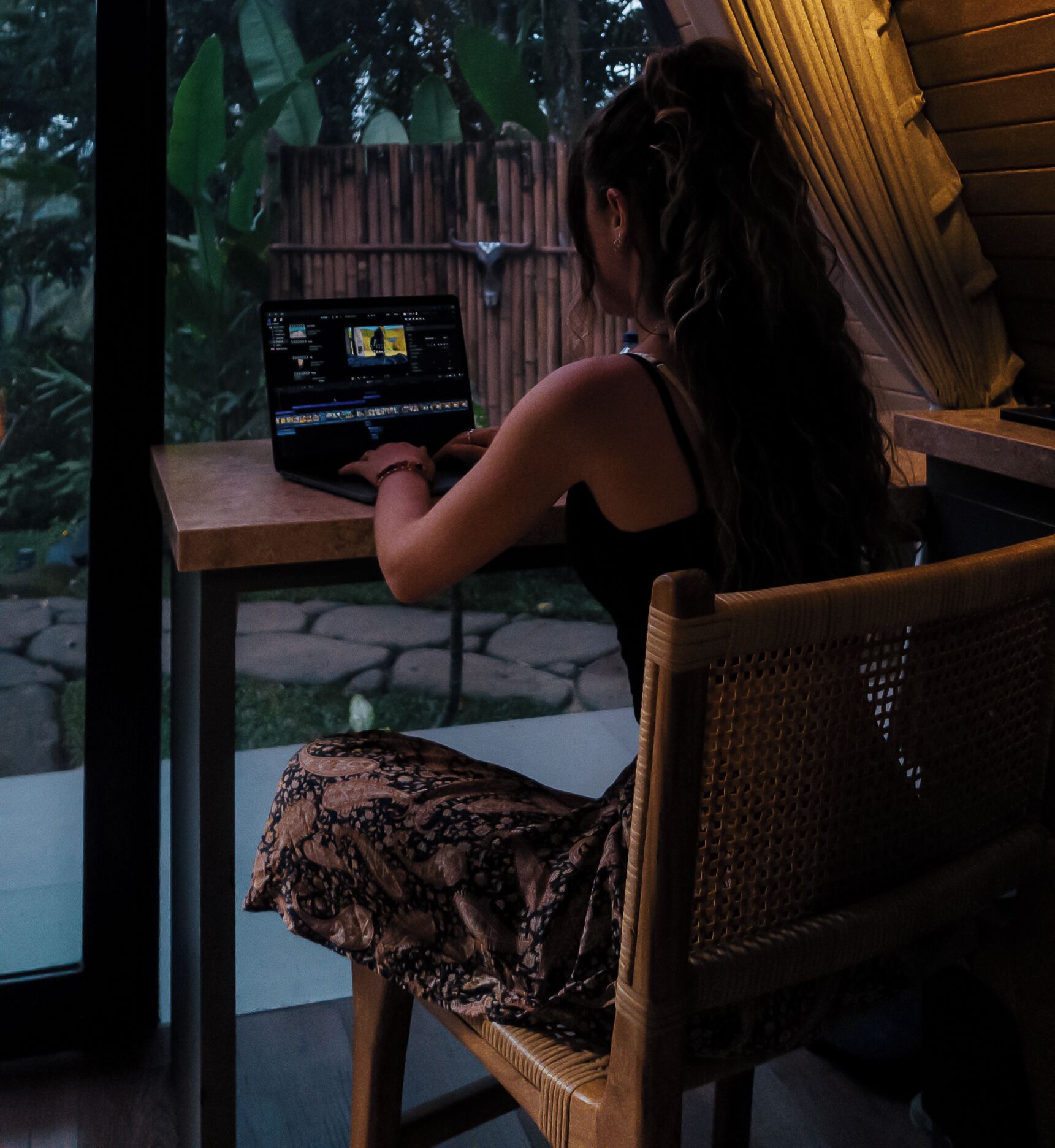 Una persona usando una computadora portátil en una mesa, con vistas a un jardín y una valla de madera.