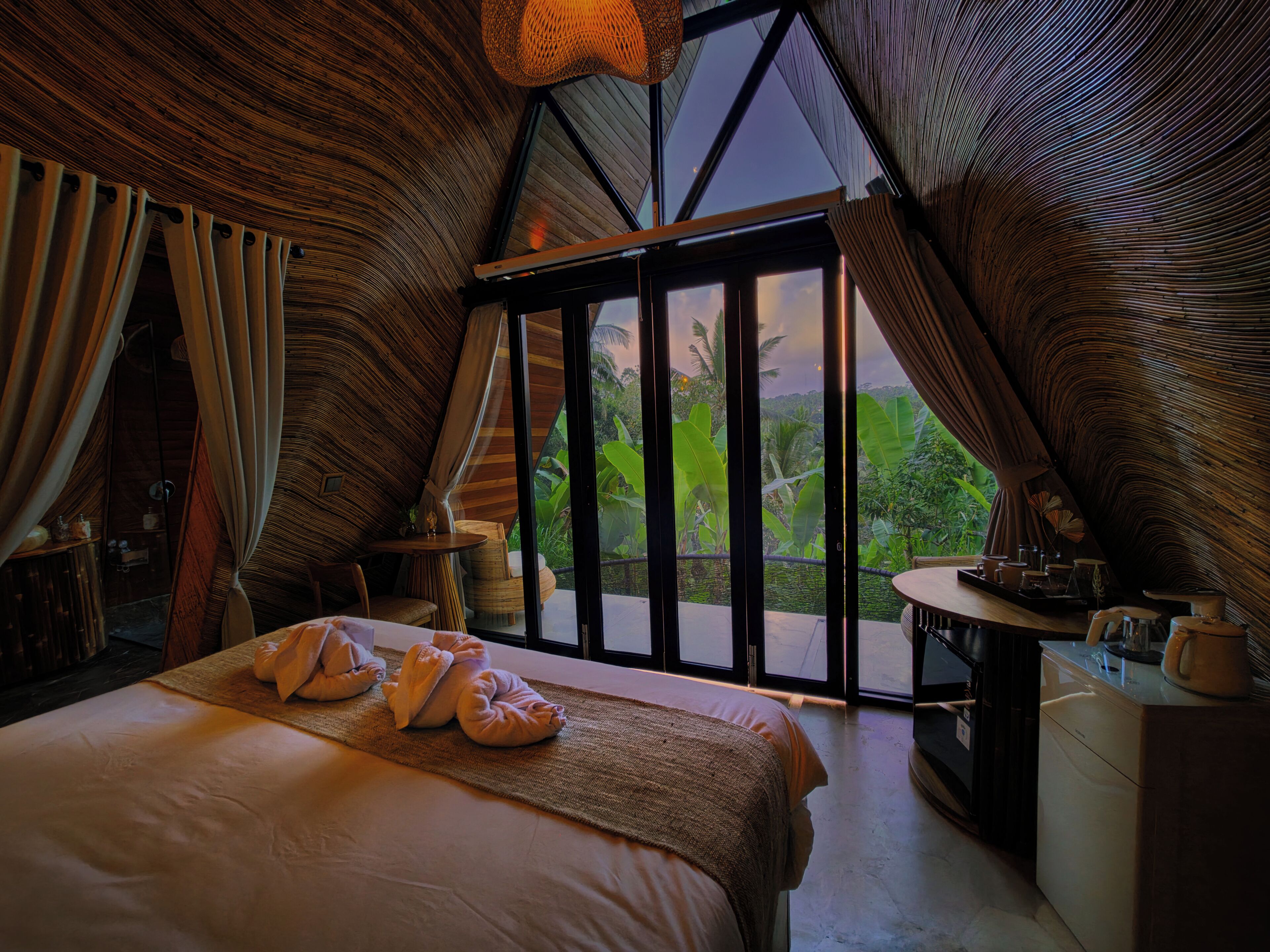 A bedroom with a large bed, a small table, and a view of lush greenery through a glass door.