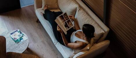 A person lying on a sofa with a magazine, a round table with a book, and a decorative vase on the floor.
