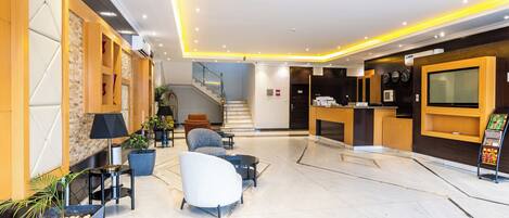 A modern lobby with seating, a reception desk, a staircase, and a TV screen.