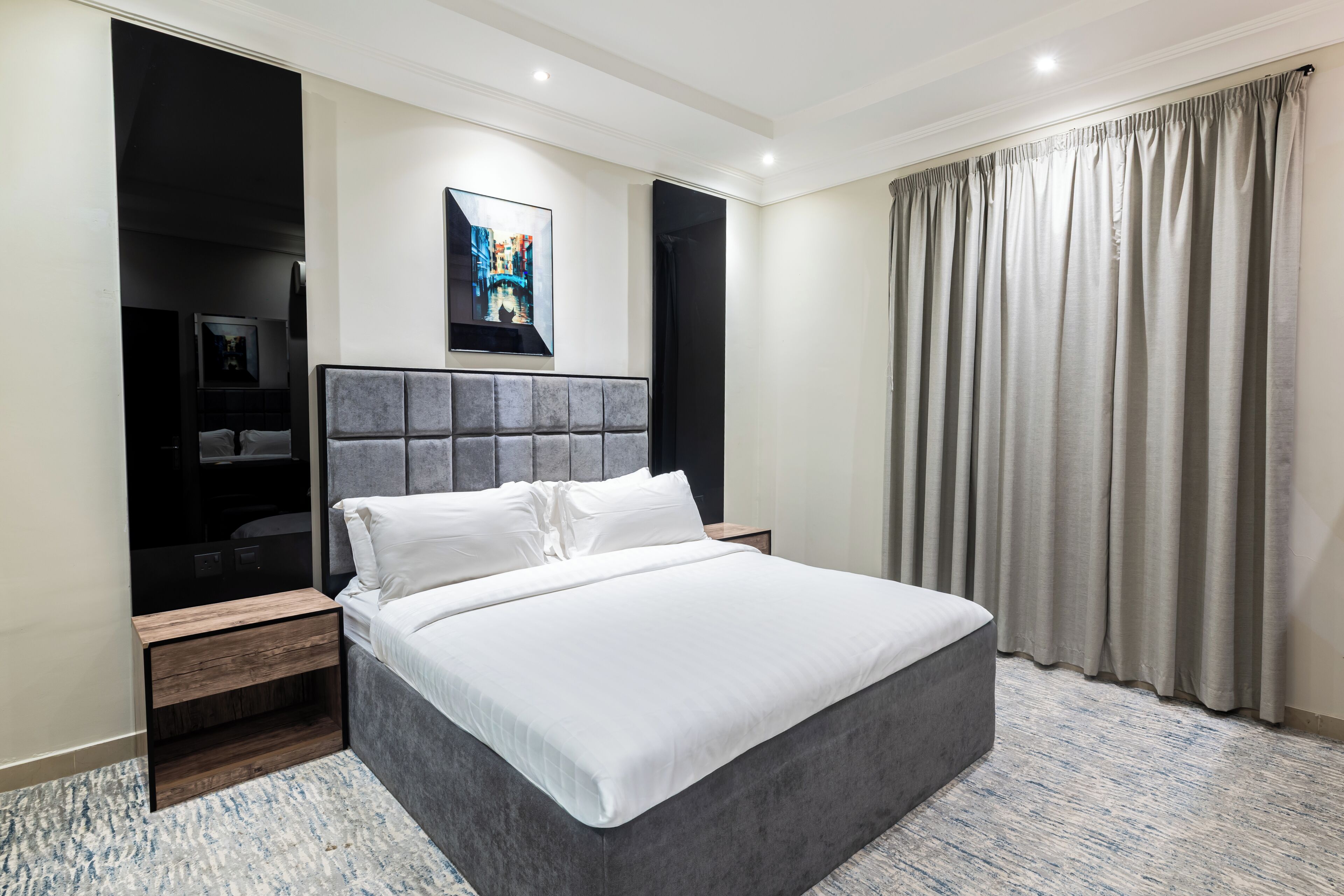 A modern hotel room with a large bed, a wardrobe, a small desk, and a painting on the wall.