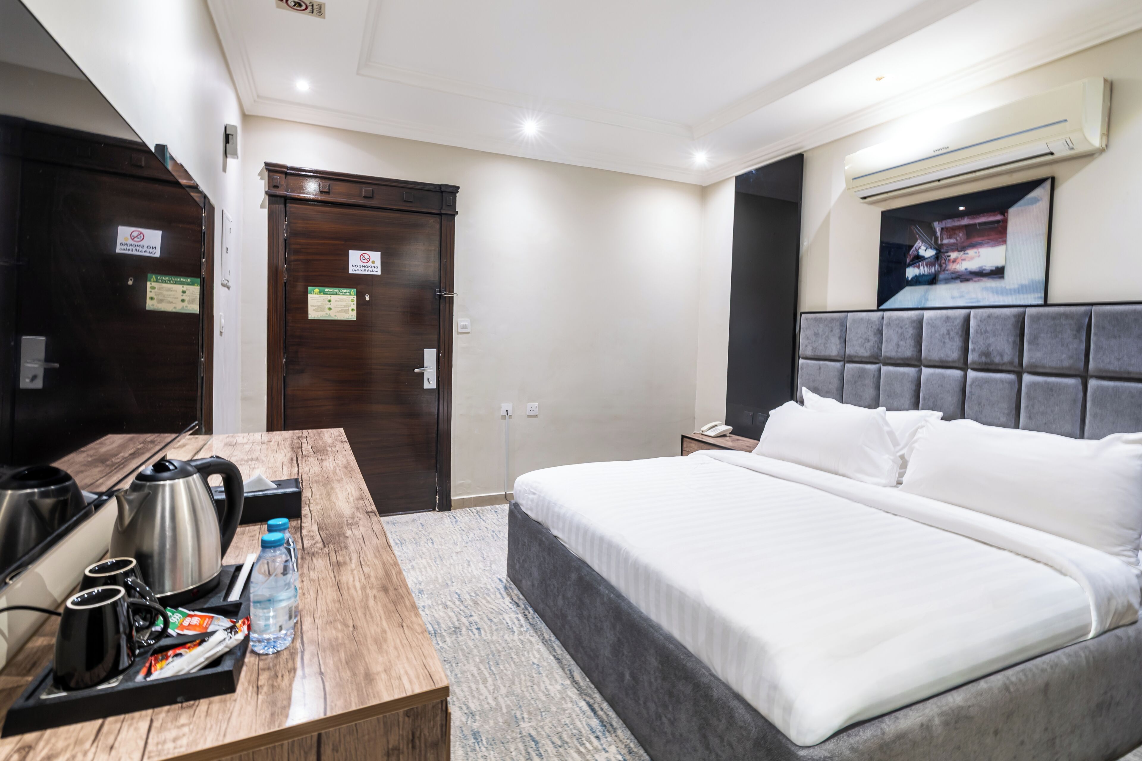 A hotel room with a bed, a desk with a kettle, cups, and a water bottle.