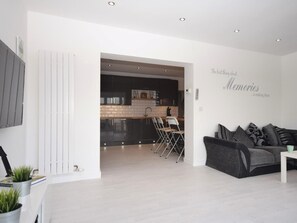 A modern living room with a grey sofa, a coffee table, and a kitchen in the background. - 4 Bed in Bideford (oc-55573) (Appledore)