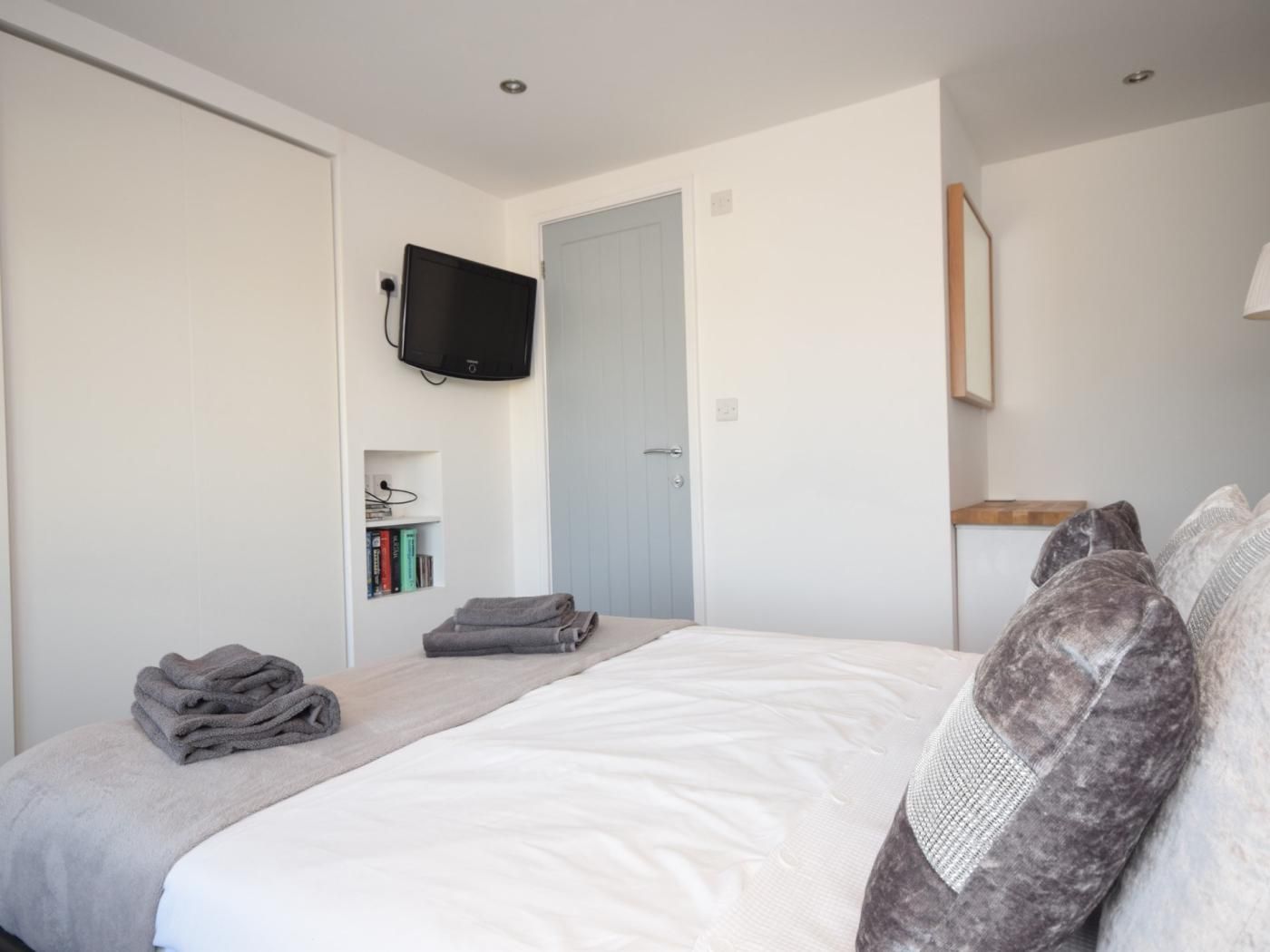 A bedroom with a bed, a television mounted on the wall, a door, and a bedside table with a lamp.