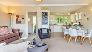 Un amplio salón con sofá, una mesa de centro y una mesa de comedor con sillas. Se observa una cocina al fondo con armarios blancos y una ventana.