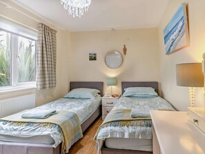 A bedroom with two beds, a window with curtains, a chandelier, a round mirror, and a painting on the wall. - 5 Bed in Tenby (oc-56683) (Penally)
