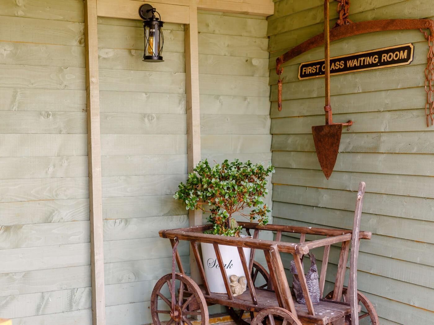 A rustic wooden structure with a sign reading 'FIRST CLASS WAITING ROOM,' a lantern, a shovel, and a cart with a plant.