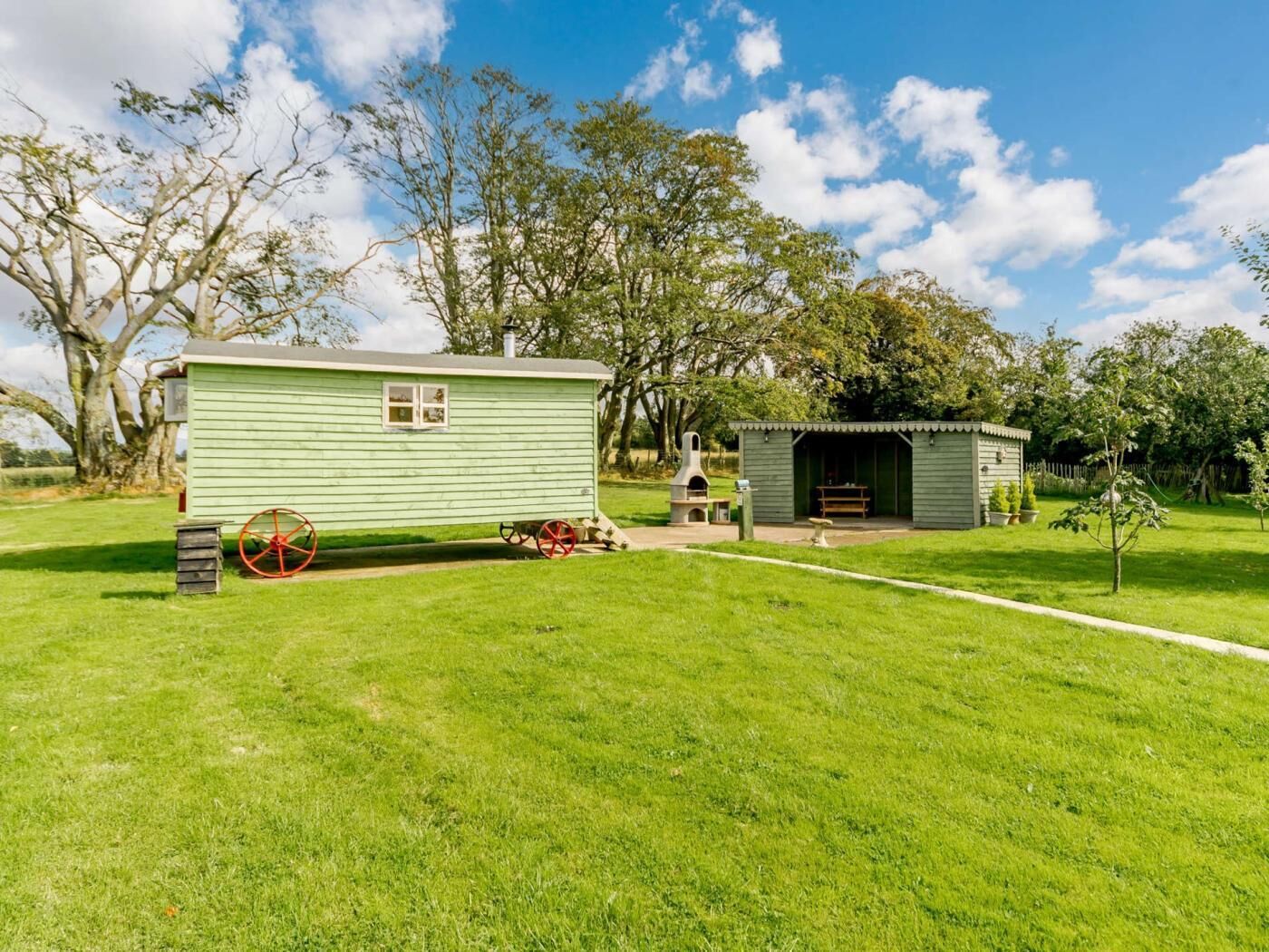 A green mobile home with a porch, a small shed, and a barbecue area in a grassy yard.