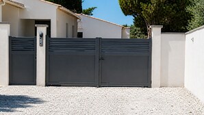 Ein verschlossener Eingang mit einem Metallgitter, einer Schotterauffahrt und einem Gebäude mit sandfarbener Fassade und einem Ziegeldach.