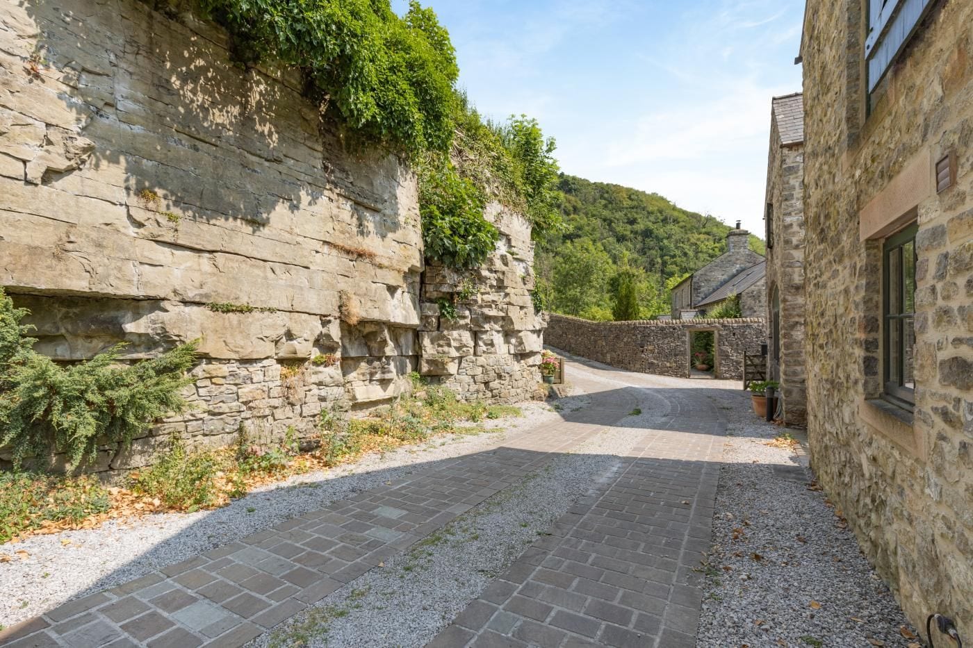 A stone building with a paved courtyard, a stone wall with vegetation, and a clear sky.