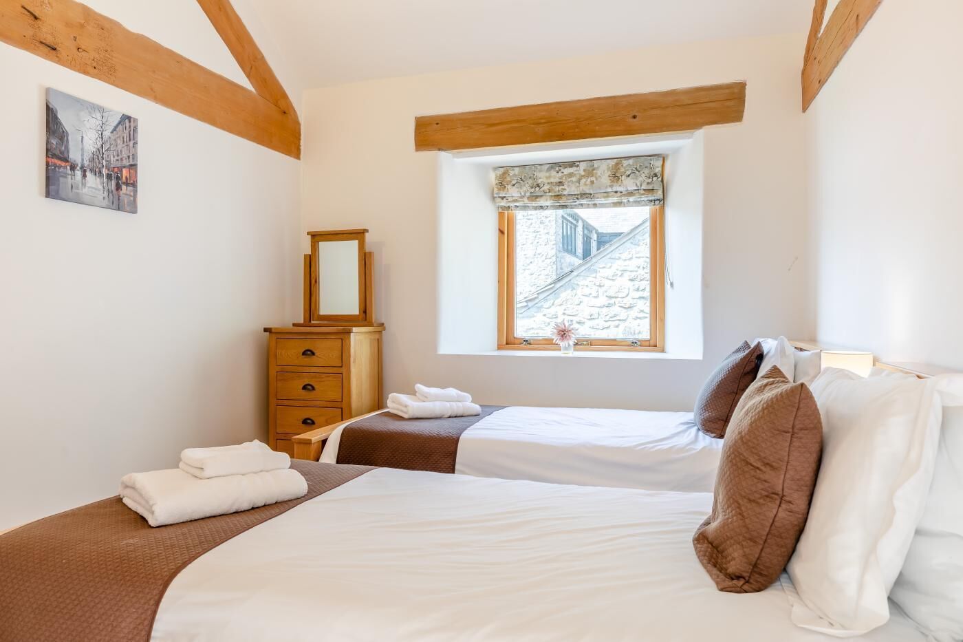 Une chambre avec deux lits, une commode, une fenêtre donnant sur un bâtiment en pierre et un tableau accroché au mur.