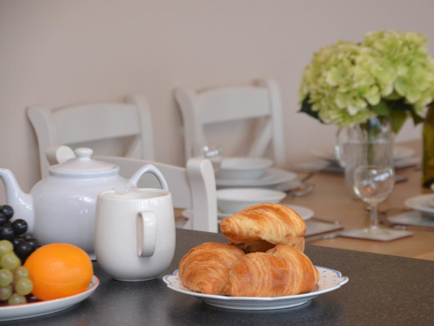 En frukostuppdukning med en tekanna, croissanter, vindruvor och en apelsin på ett bord.