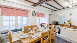 A dining area with a wooden table set for four, a kitchen in the background, and a 'WELCOME' sign on the window. - 3 Bed in Ilfracombe (oc-57795) (Ilfracombe)