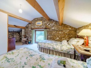 Une chambre avec deux lits, un mur en pierre, des poutres en bois, une fenêtre avec des rideaux et une table de chevet avec une lampe.