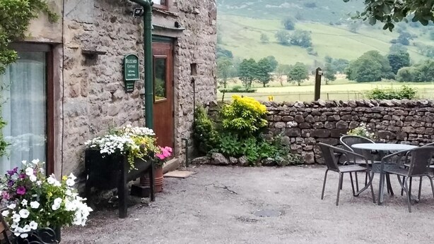 A stone building with a sign reading 'Buzzard Cottage.' There is a bench and outdoor seating area with a table and chairs. - 2 Bed in Sedbergh (oc-57641) (Dent)