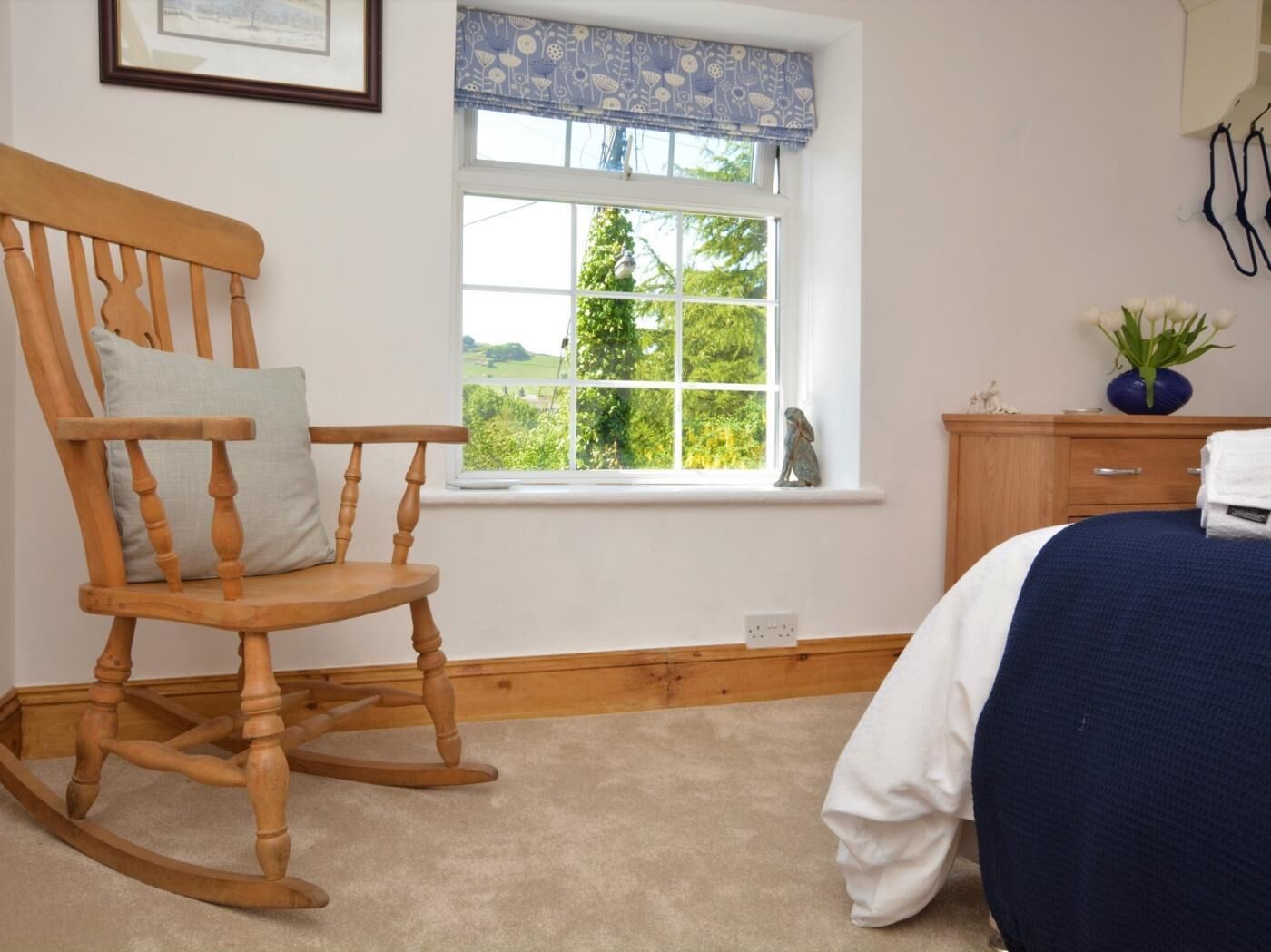 Une chambre avec un fauteuil à bascule, un lit recouvert d’une couverture bleue, une fenêtre donnant sur un paysage verdoyant et une petite armoire ornée d’une plante.
