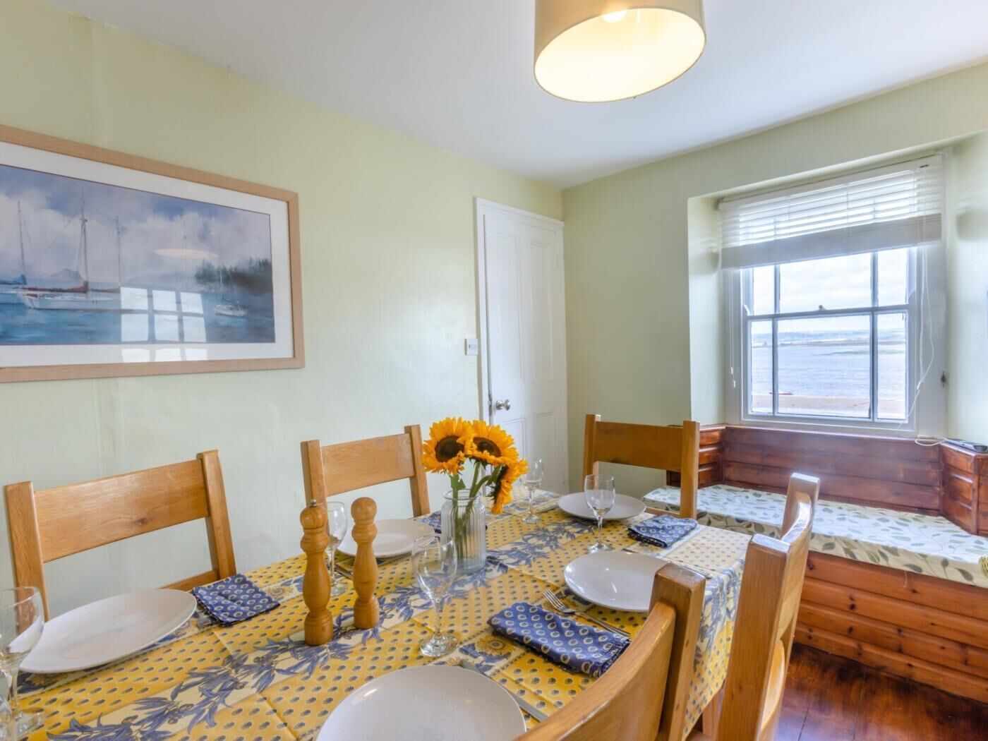 Un comedor con una mesa dispuesta para cuatro personas, un cuadro en la pared y una ventana con vista al mar.