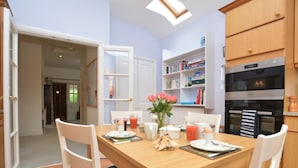 Un coin repas avec une table en bois, des chaises blanches, une bibliothèque et une cuisine équipée d’un four.