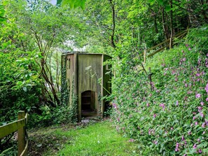 A rustic wooden structure with a door, surrounded by lush greenery and flowering plants. - 3 Bed in Bideford (oc-60643) (Buck's Mills)
