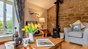A cozy living room with a stone wall, a fireplace, a sofa, a coffee table with breakfast items, and a window with curtains. - 2 Bed in Morpeth (oc-61267) (Rothbury)