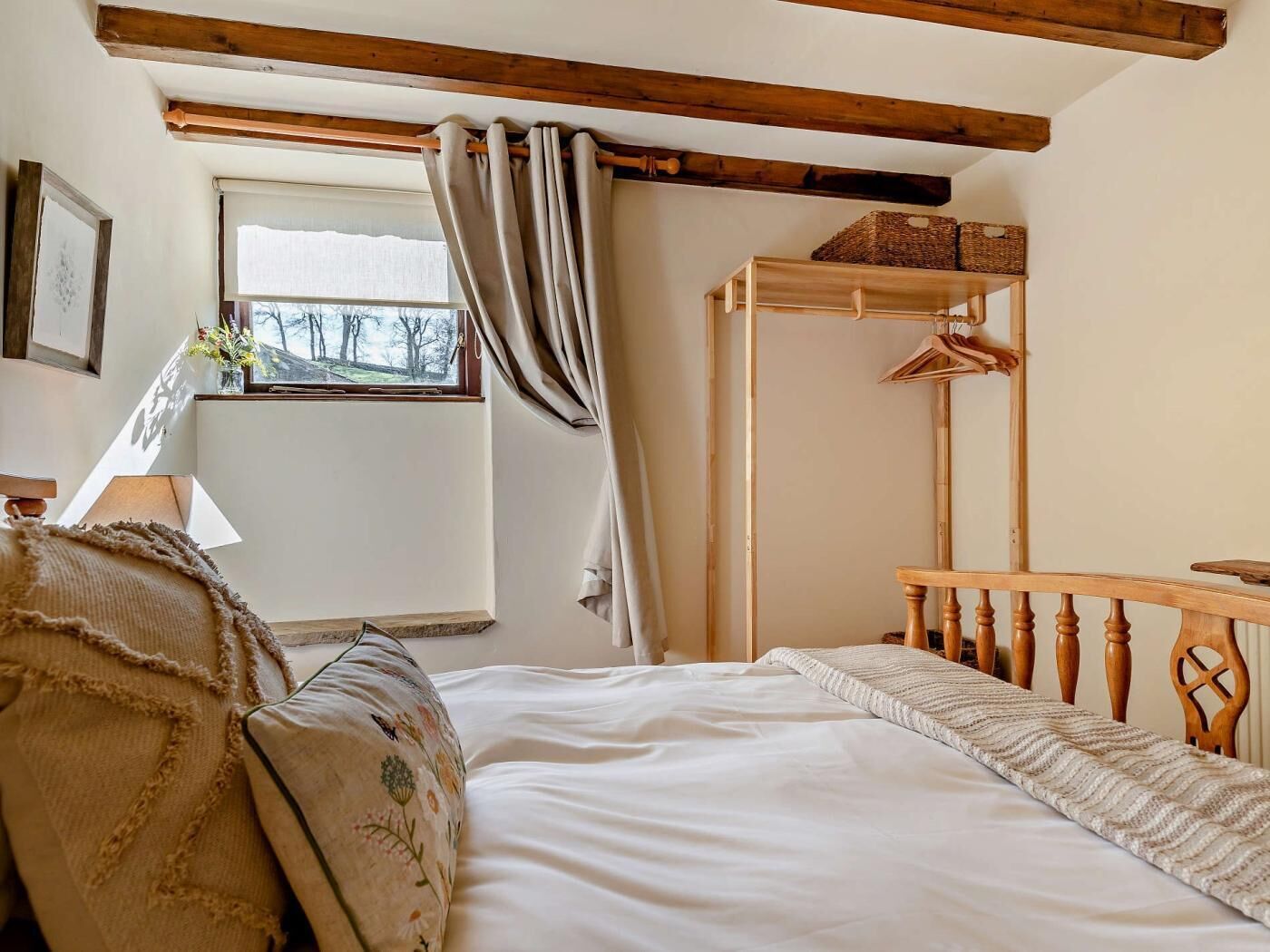 A bedroom with a bed, a wooden headboard, a window with curtains, a bedside table, and a hanging clothes rack.
