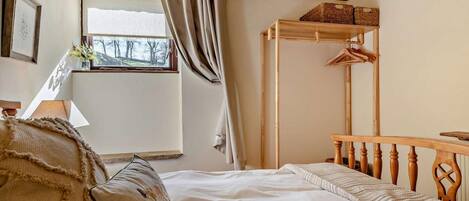 A bedroom with a bed, a wooden headboard, a window with curtains, a bedside table, and a hanging clothes rack.
