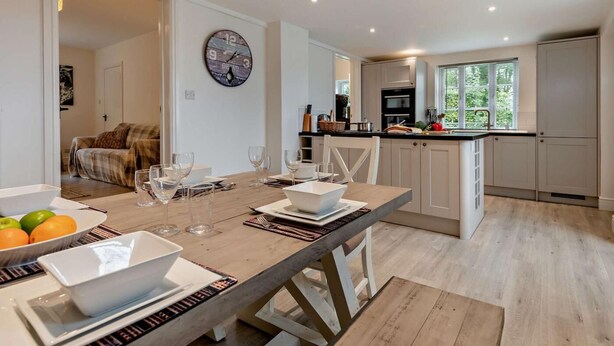 Une cuisine moderne avec des meubles blancs, un coin repas doté d’une table en bois pour quatre personnes, et une grande horloge au mur.